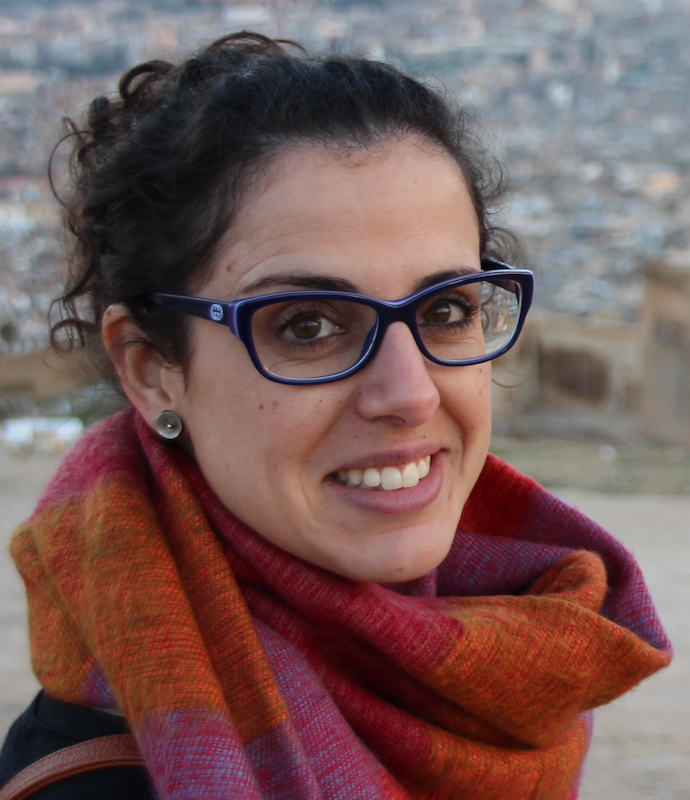 Colour headshot of a white-presenting woman smimling and looking into the camera. She has dark hair, glasses and wears a brightly coloured neck scarf.