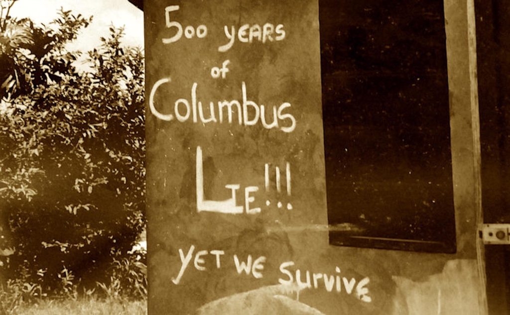Sepia photo of graffitti on a wall reading 500 years of Columbus Lie!!! yet we survive