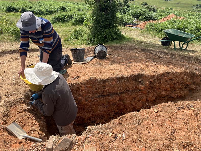 Colour photograph of two people wearing sun hats, one standing in an archaeological trench, the oter knelling alongside.