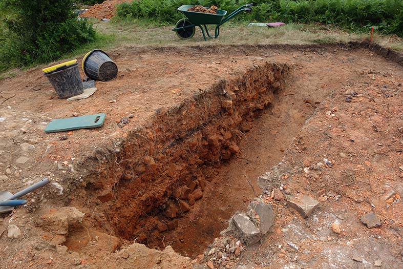 Colour photograph of a rectangular archaeological trench cutting through brick-red soil and layers of broken bricks. A thin dark black line is visible amongst the layers.