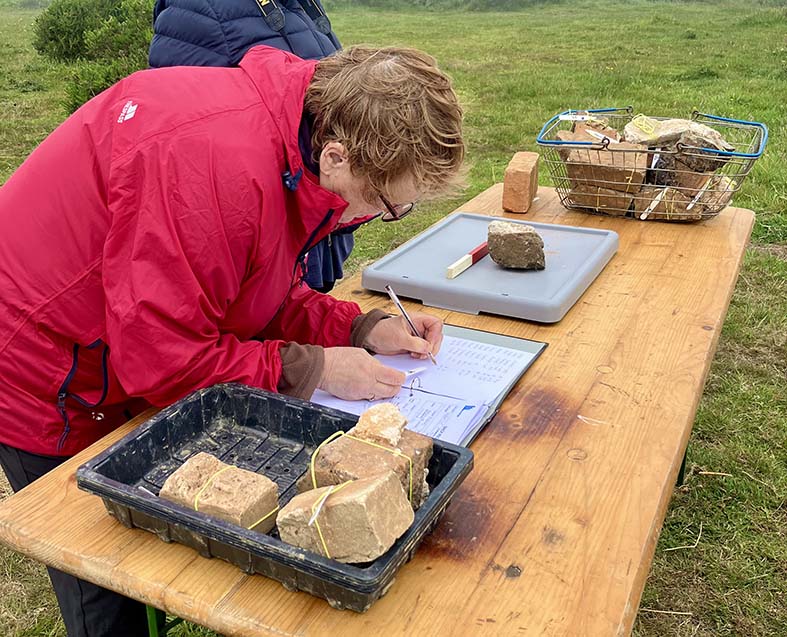 COlour photograph of a person wearing a red coat leaning over a table out of doors, writing on a record sheet with trays of bricks on the table.