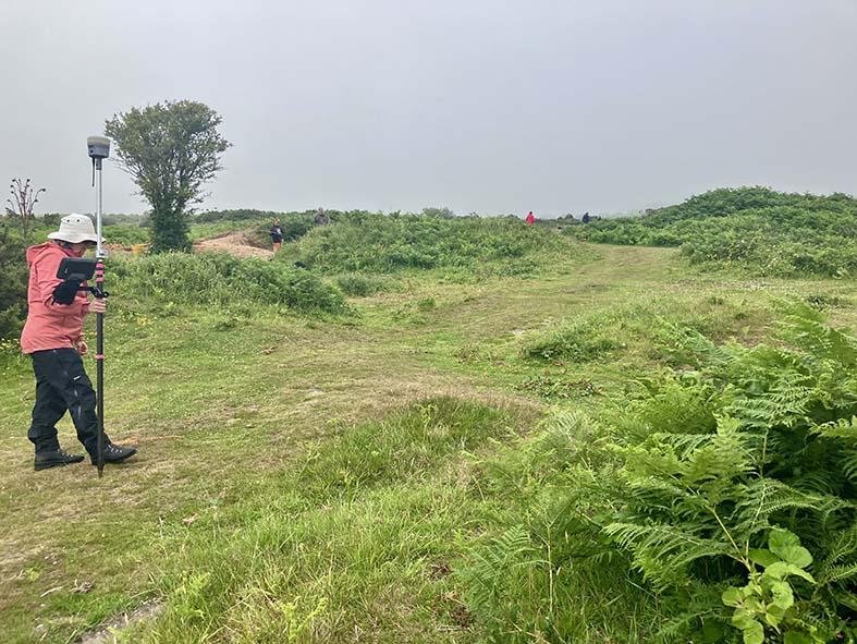 Colour photograph of a person ewaring a white sun hat, red coat and black trousers walking over a grassy area with braken growing nearby.They are carrying a tall pole with a GPS receiver fixed on the top.