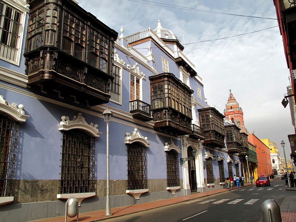 Colour photo of an ornate building of more than 9 bays with grills on ground floor windows and oriel windows on the first floor, along a street front.