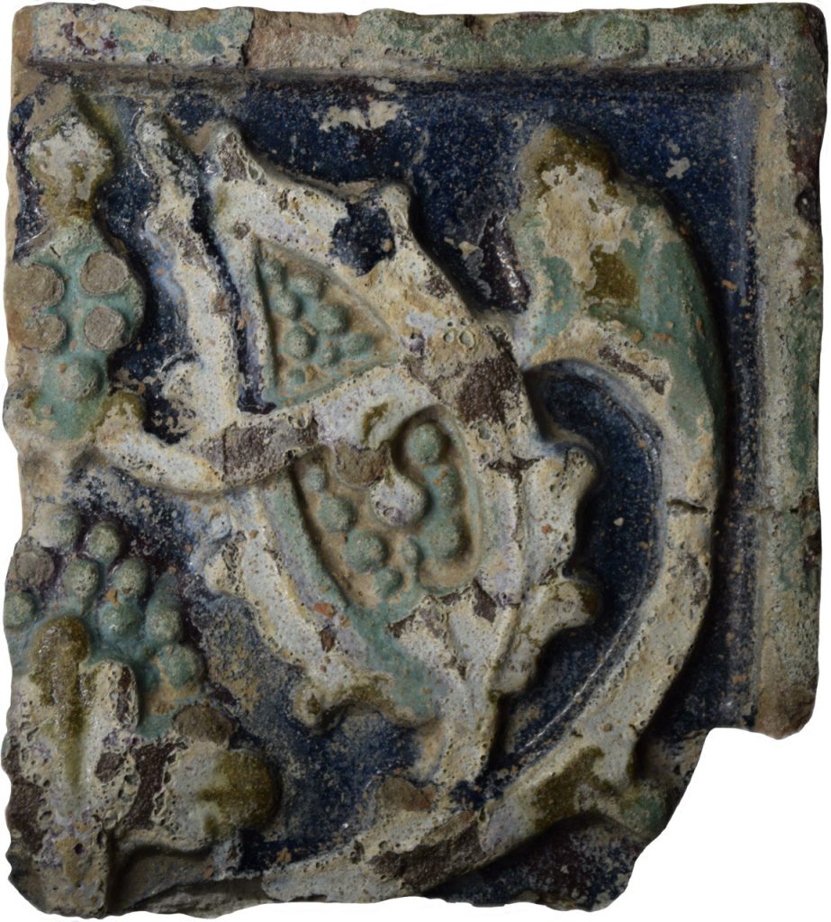 Colour photo of a floral patterned stove tile in black, grey, teal and yellow glazes.