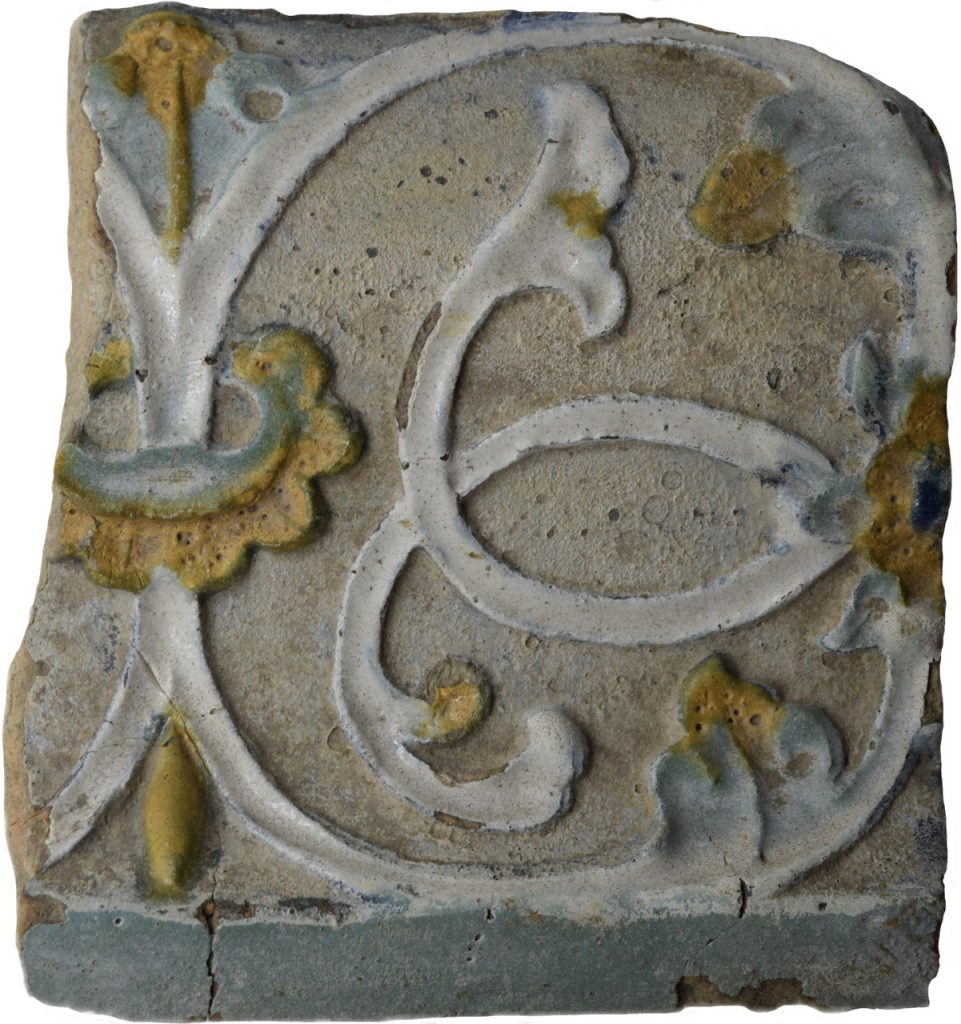 Colour photo of a floral patterned stove tile in black, grey, teal and yellow glazes.