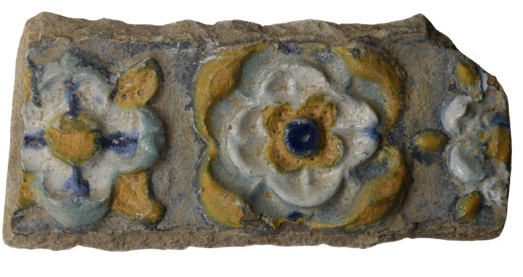 Colour photo of a floral patterned stove tile in black, grey, teal and yellow glazes.