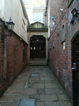 Colour photo of a passage between brick buldings laid to stone paving slabs, leading to a broad doorway with a pale stone surround.