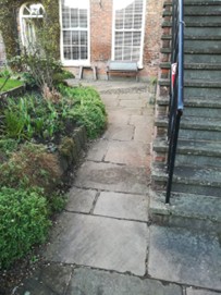 Colour photo of a pathway in a garden laid to stone paving slabs, with greenery to left and a stone staircase to right.