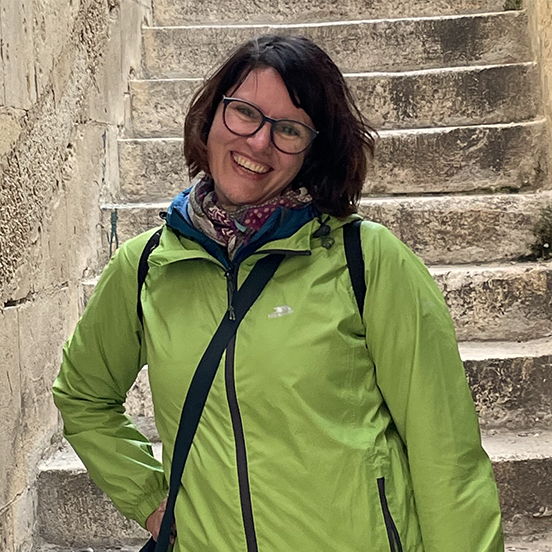 Colour photo of a white presenting woman with dark shoulder length hair, glasses and a lime green top smiling at the camera.