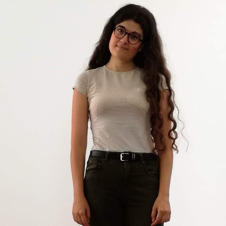 Colour three-quarter photo of a young woman with long dark hair, wearing glasses, a pale short-sleeved top and dark trousers with a belt.