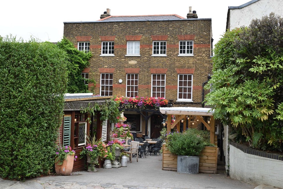 Colour photo of a three storey yellow and red brick pub with green hedges to either side and colourful flower pots.