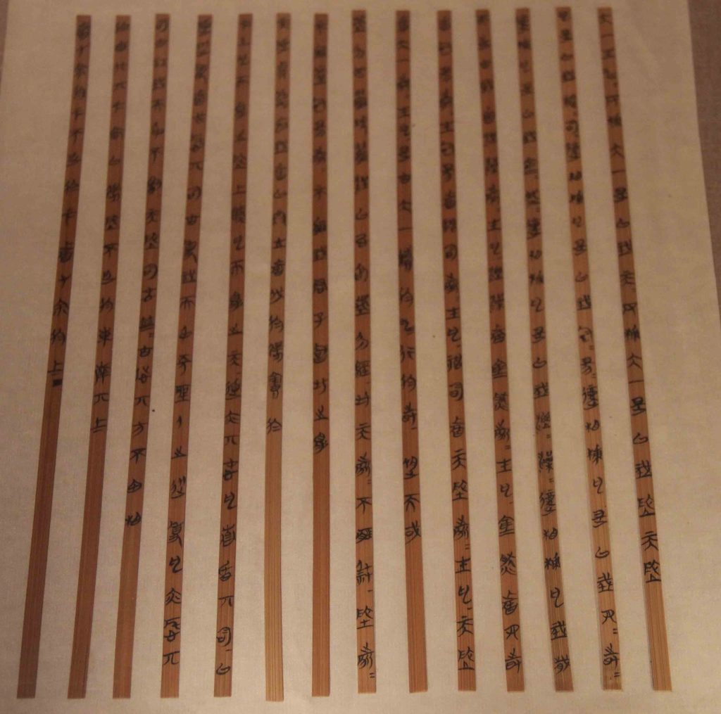 Colour photo of 14 thin brown bamboo strips side by side with characters written on each face in black ink.