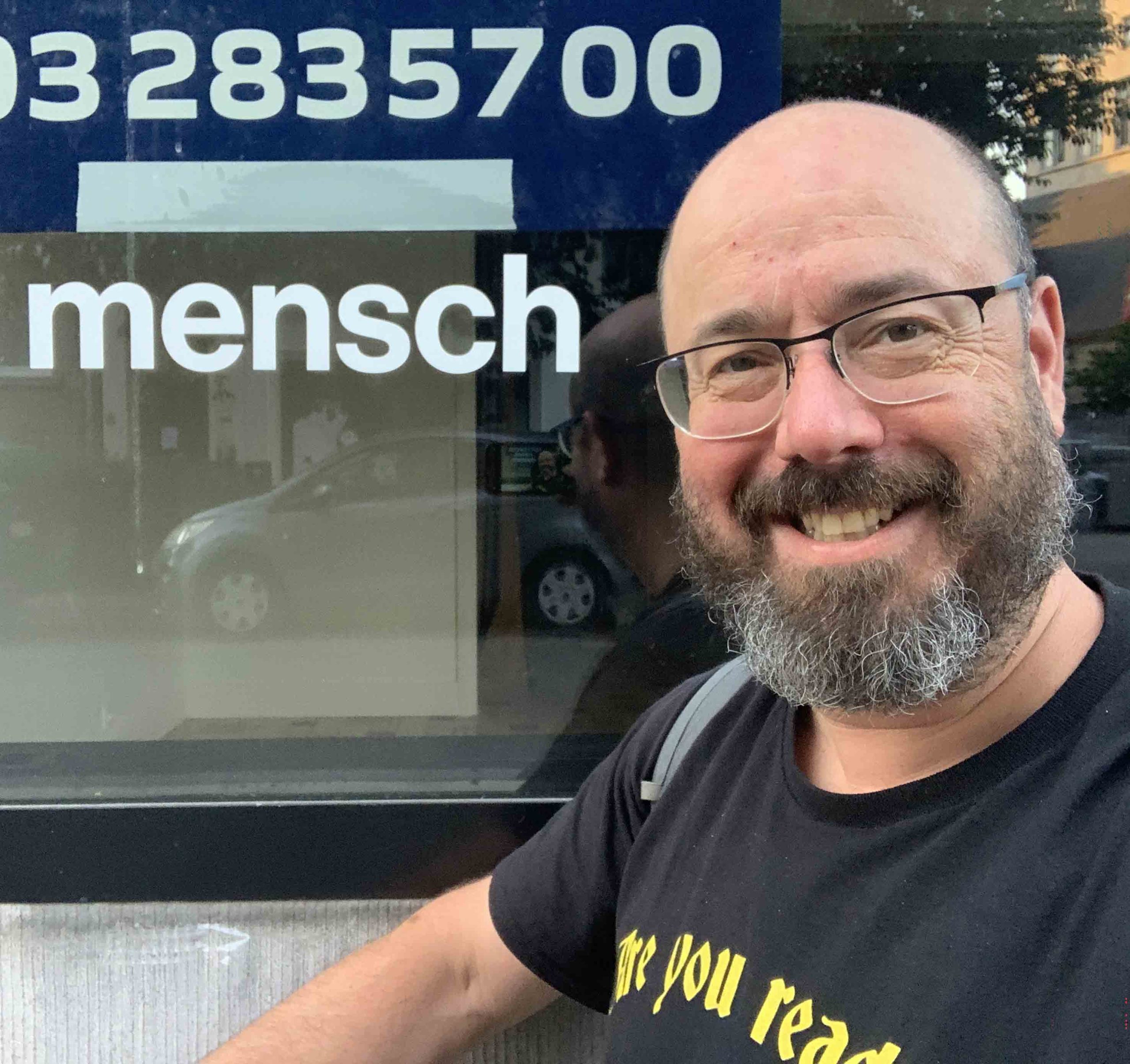 Colour photo: a headshot of a smiling white man wearing glasses, dark t-shirt and a beard standing in front of a window. A string of numbers and the word mensch are printed in pale blue on the window glass.