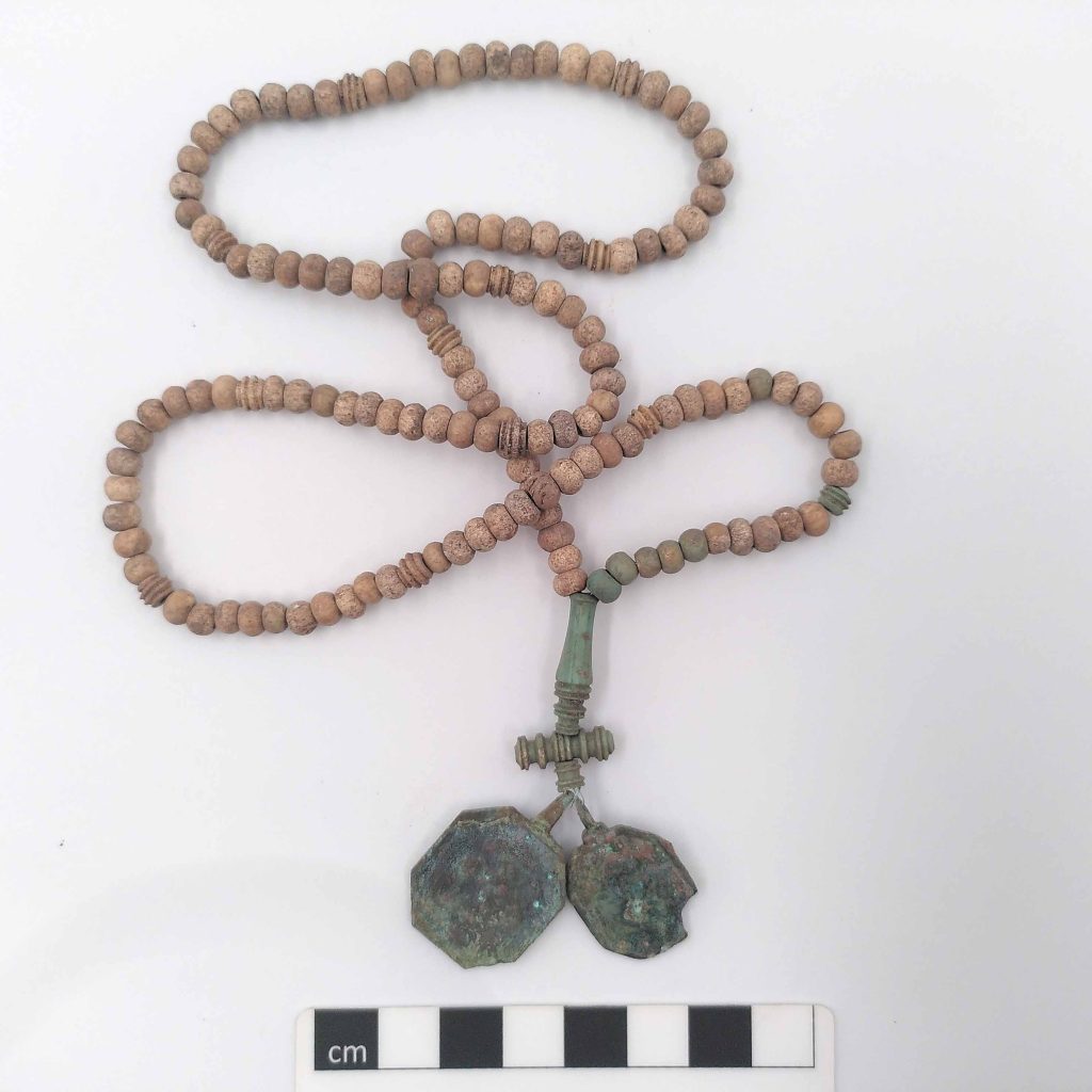 A string of small pale brown circular beads, some smooth and some ribbed, ending with green metal beads in a cross shape and two small octagonal metal discs hanging from the bottom of the cross.