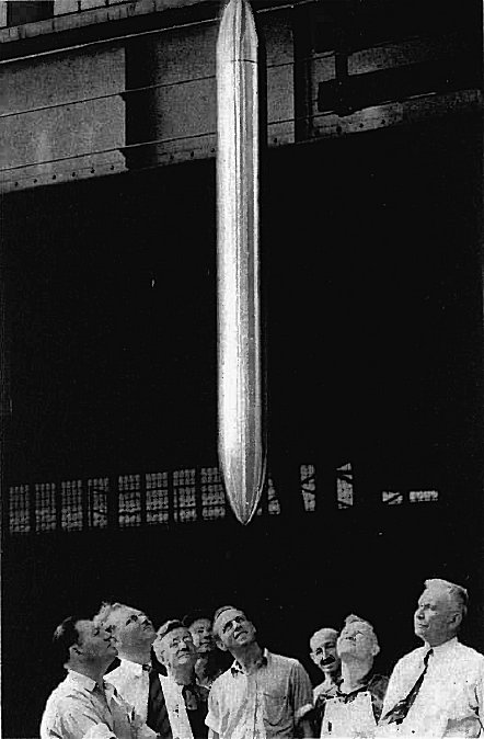 Black and white photo of a group of men looking up at a long, shiny cigar-shaped object suspended vertically above their heads.